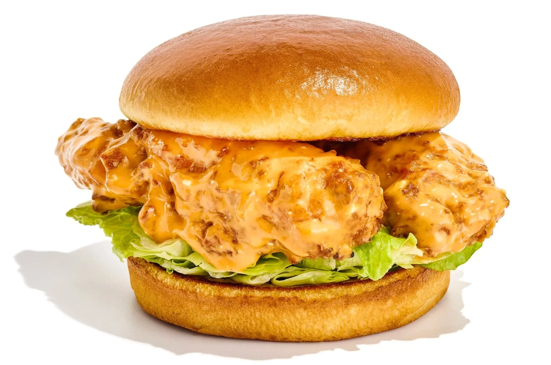 Close-up of a fried chicken sandwich with lettuce, cheese, and creamy sauce in a bun against a white background.