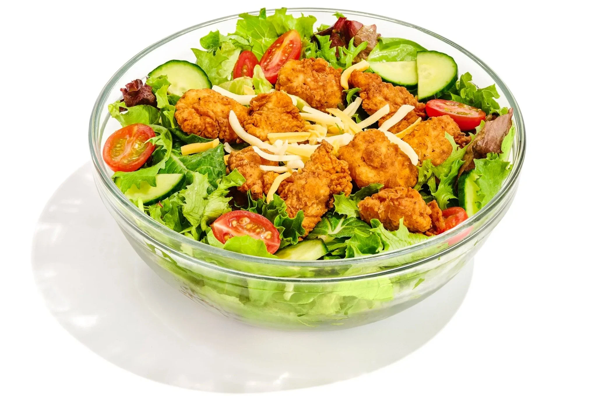 A glass bowl filled with a colorful salad that includes shredded cheese, cherry tomatoes, cucumber slices, greens, and fried chicken pieces.