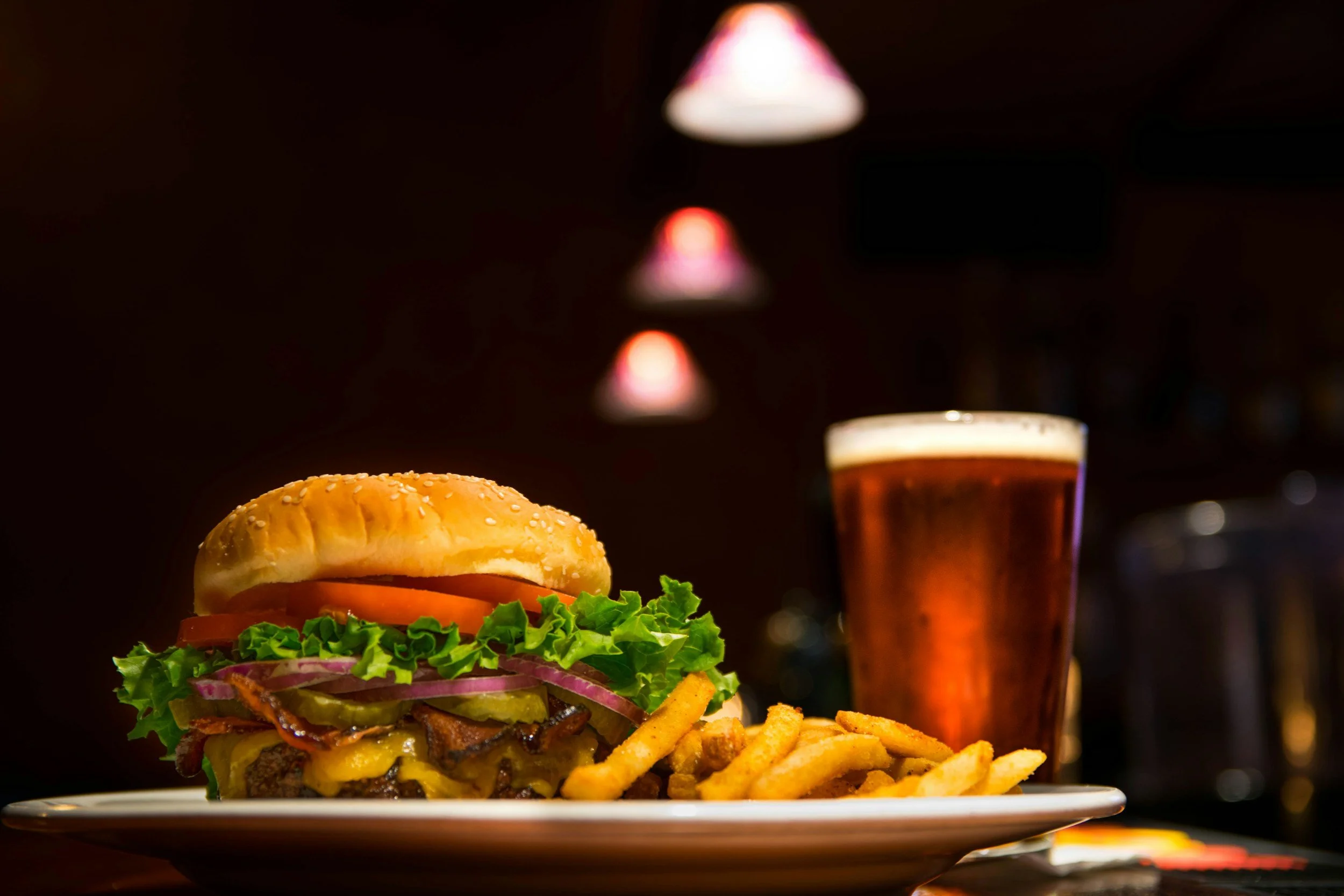 A plate with a cheeseburger containing lettuce, tomato, pickles, onions, bacon, and cheddar cheese, accompanied by curly fries, with a pint of beer and bar lights in the background.