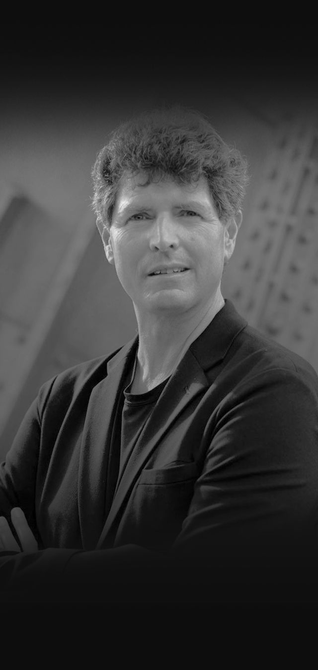 Black and white portrait of a man with curly hair wearing a blazer and t-shirt, looking at the camera with a serious expression.