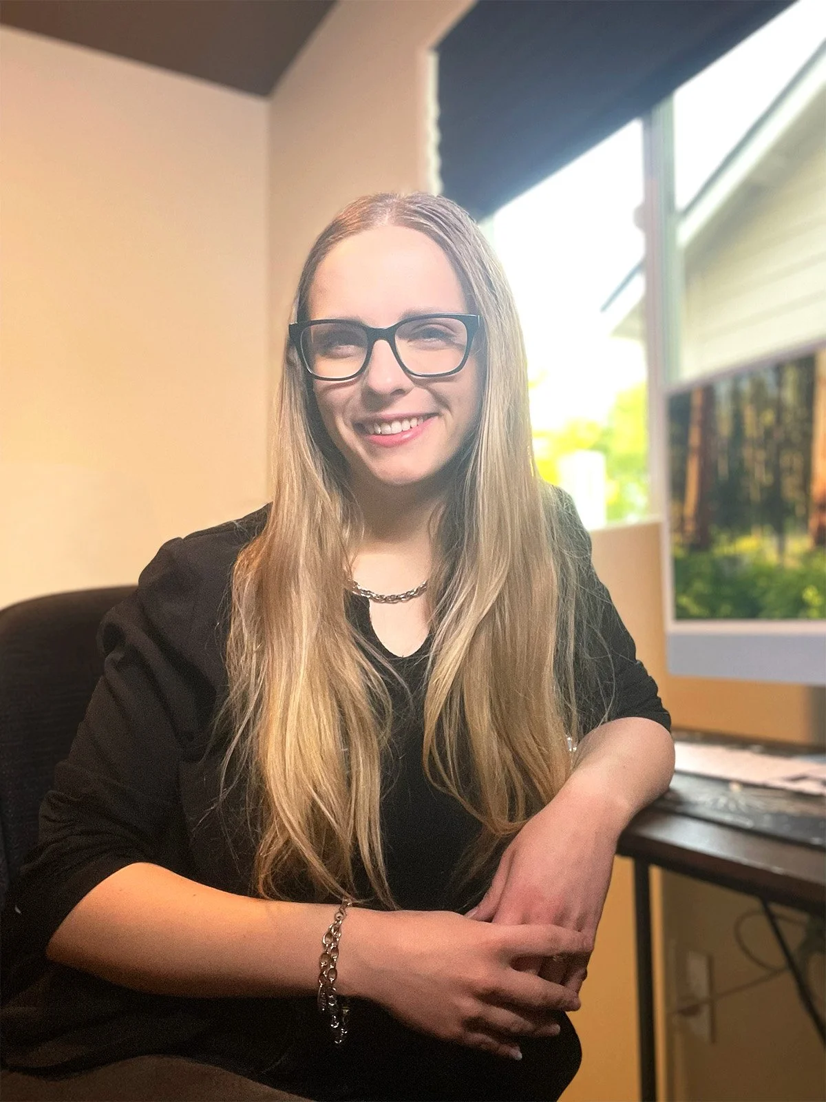 A woman with long blonde hair wearing glasses, a black top, a silver necklace, and a bracelet sitting at a desk with a window showing a green outdoor scene in the background.