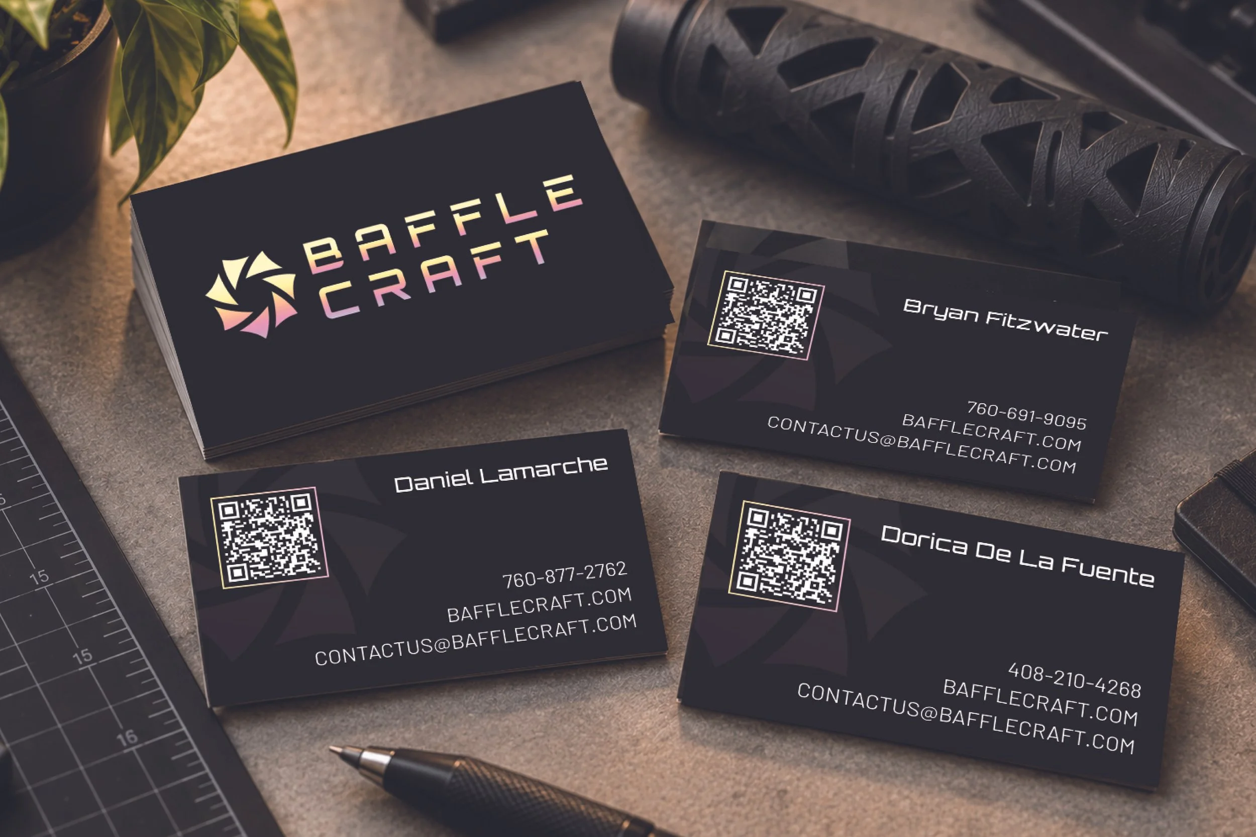 A stack of black business cards with a colorful logo and white text, a QR code, and contact information for Baffle Craft, with some business cards laid out on a gray surface alongside a potted plant, a utility knife, and a black textured roller.