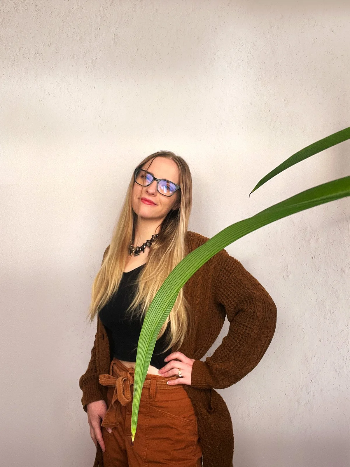 A woman with long blonde hair, glasses, and a confident smile, posing with her hand on her hip in front of a plain white wall. She is wearing a black top, a brown cardigan, and brown high-waisted pants with a tied belt, accessorized with a black beaded necklace.