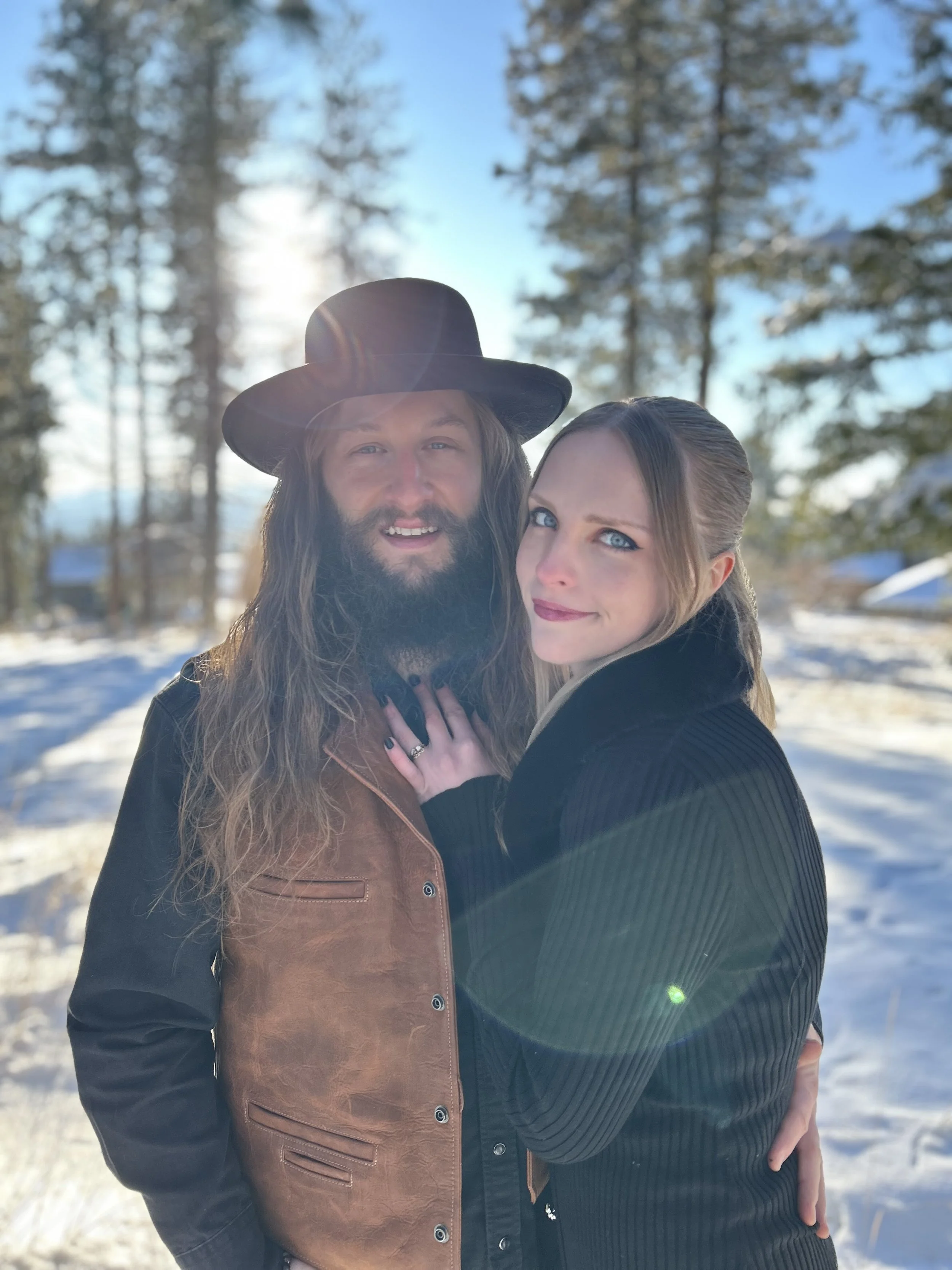 A man with long hair and beard wearing a black hat and a brown leather jacket, and a woman with blue eyes and long hair wearing a black sweater, standing outdoors in a snowy woodland area with trees and a clear blue sky in the background, illuminated by sunlight.
