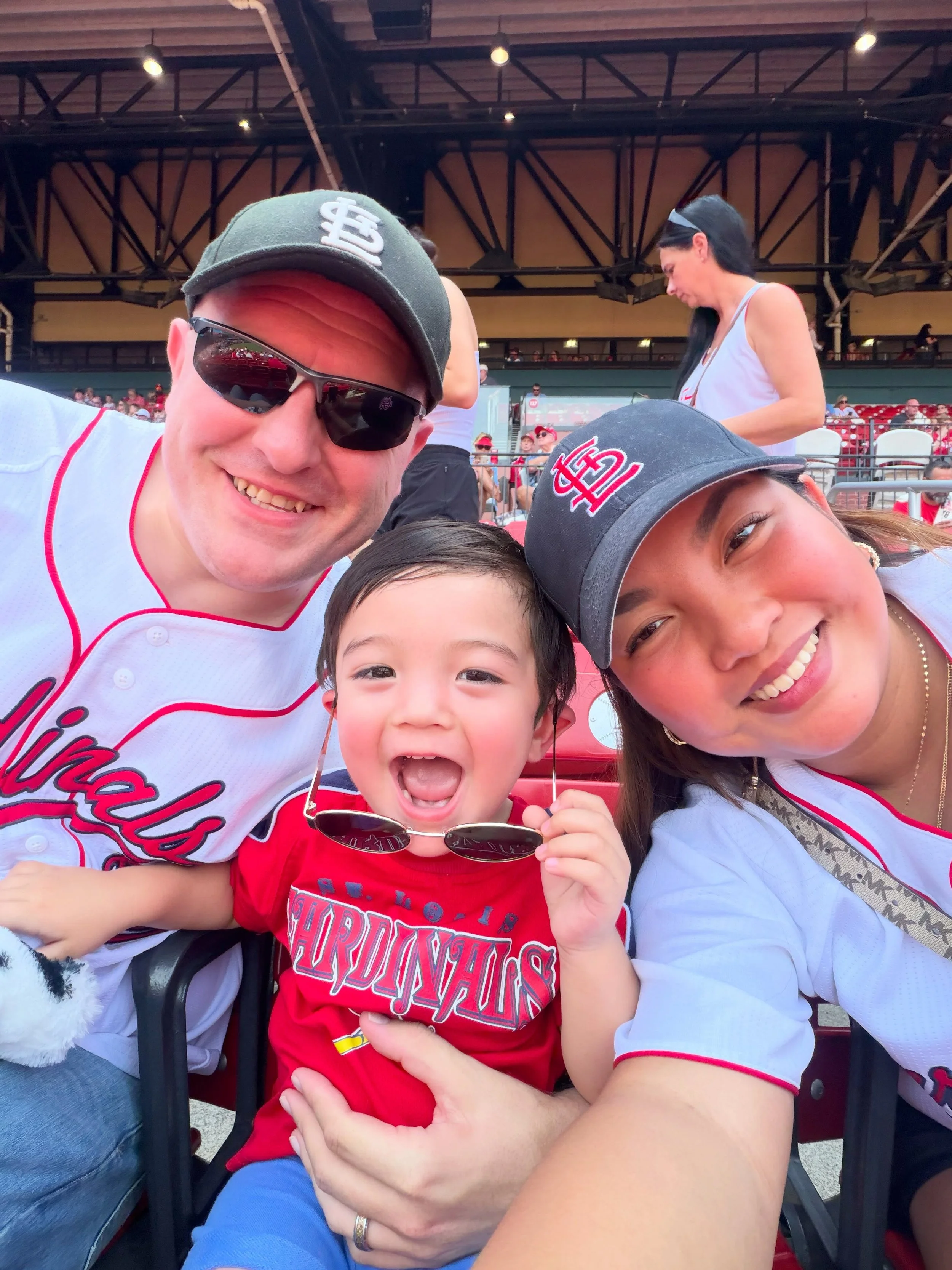 A group of three smiling people, including a young boy, at a baseball game. They are wearing St. Louis Cardinals hats and shirts, and appear excited. Background shows a crowded stadium with spectators.