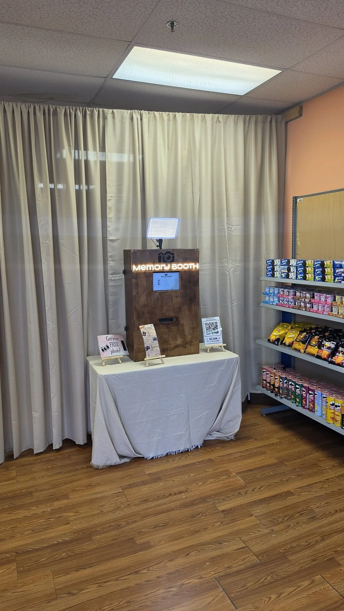 Photo of a photo booth setup with a wooden frame, a sign that reads 'Memory Booth,' a small light on top, and a tablet for taking photos, located in a room with beige curtains and shelves of snacks on the right side.