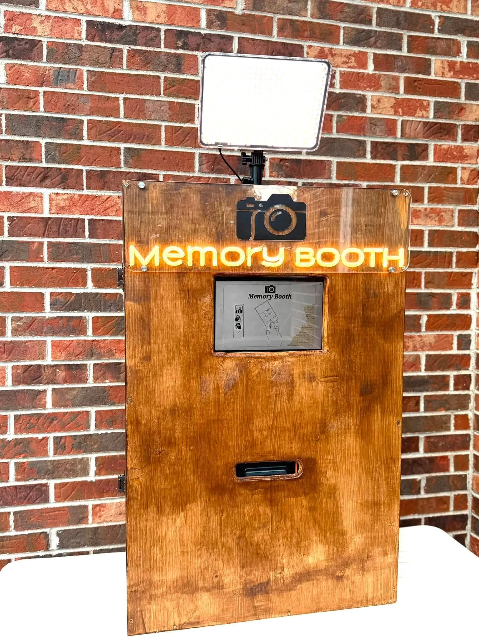 Photo of a wooden memory photo booth with a neon sign that says 'Memory BOOTH' against a red brick wall. There is a camera icon above the neon sign and a light panel on top.