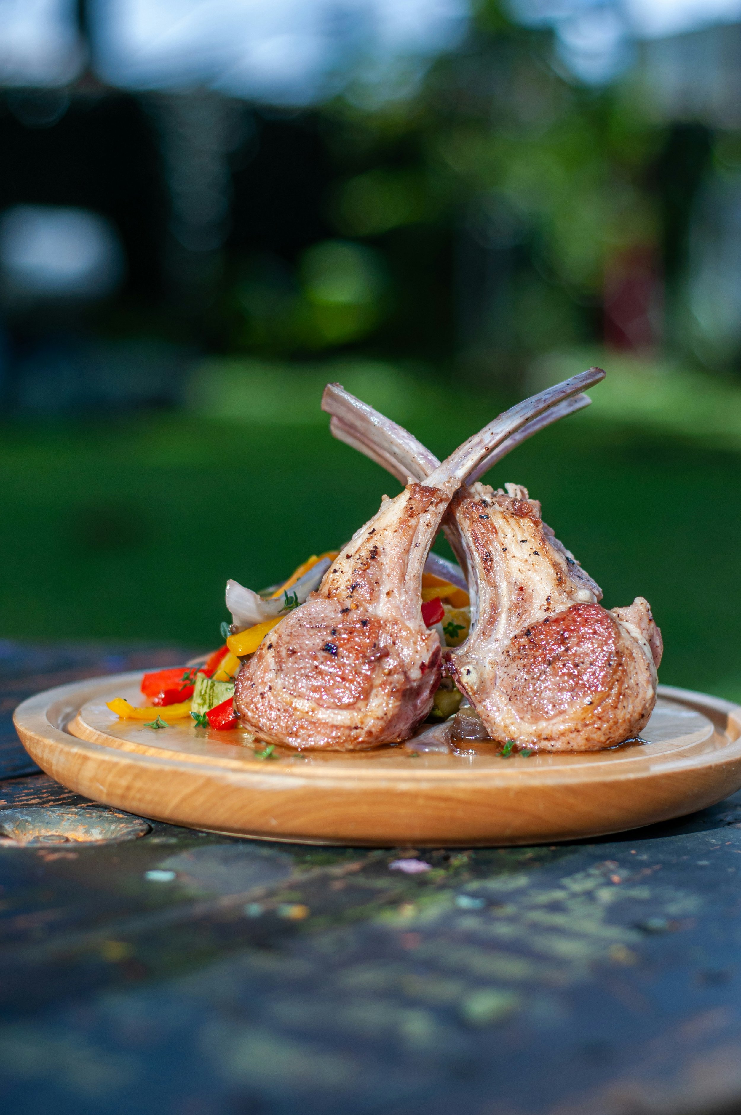 Close-up of two cooked lamb chops served on a wooden platter with colorful vegetables underneath, set outdoors with a blurred green and blue background.