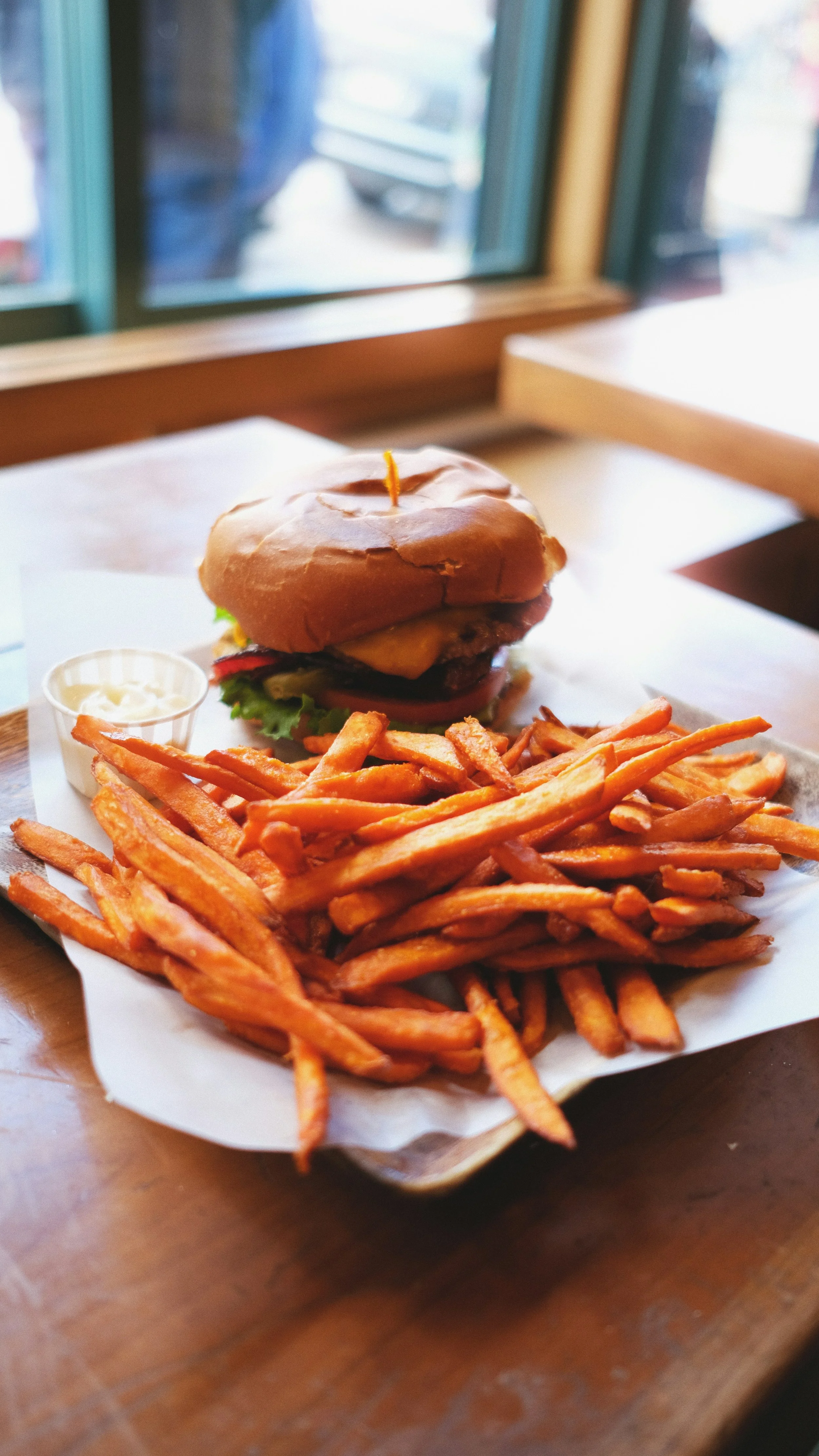 A plate with a burger, French fries, and a small cup of dipping sauce on a wooden table near a window.