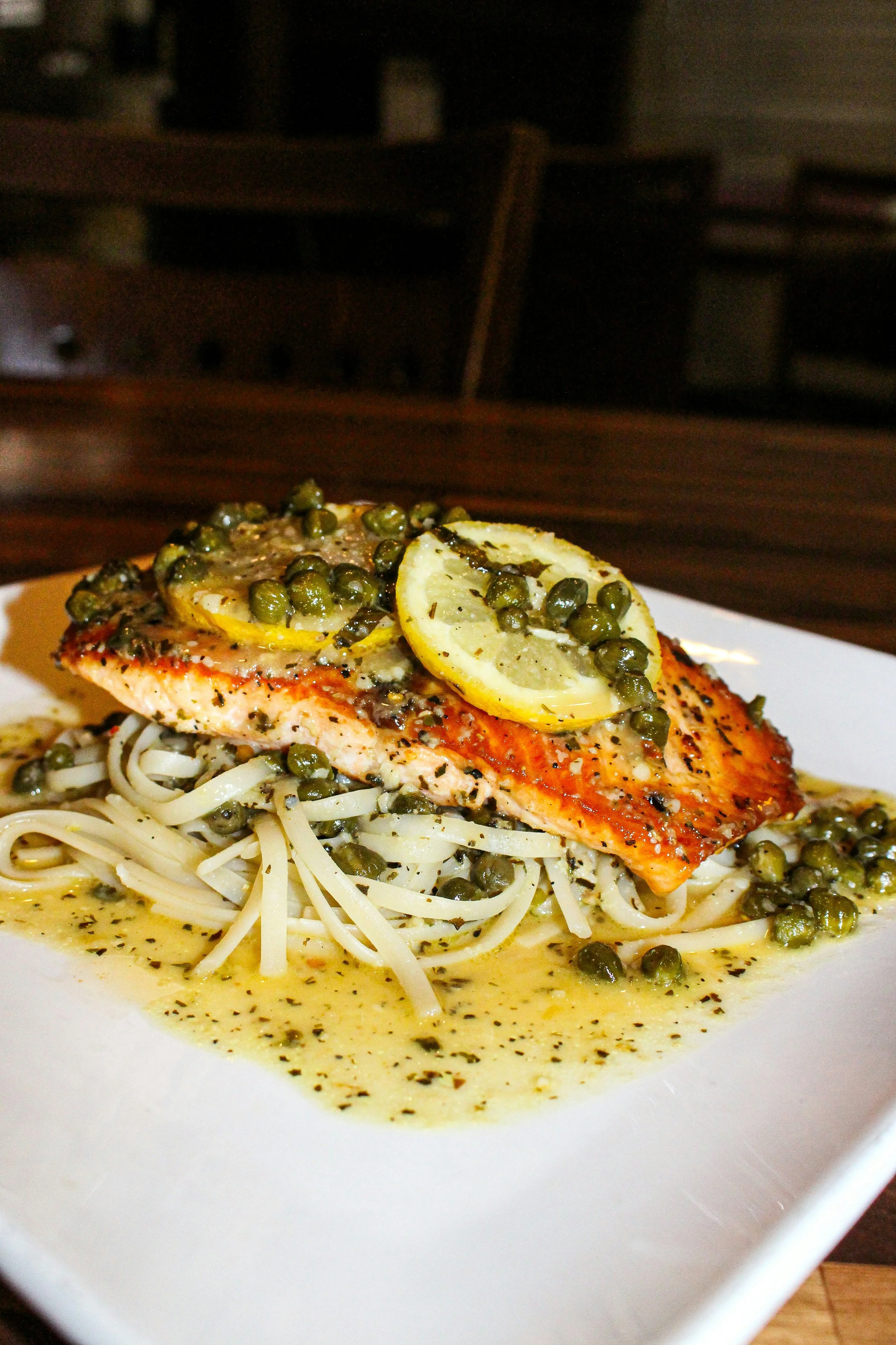 Close-up of a plate with cooked salmon topped with lemon slices and green peppercorns, served over pasta with light-colored sauce and herbs.