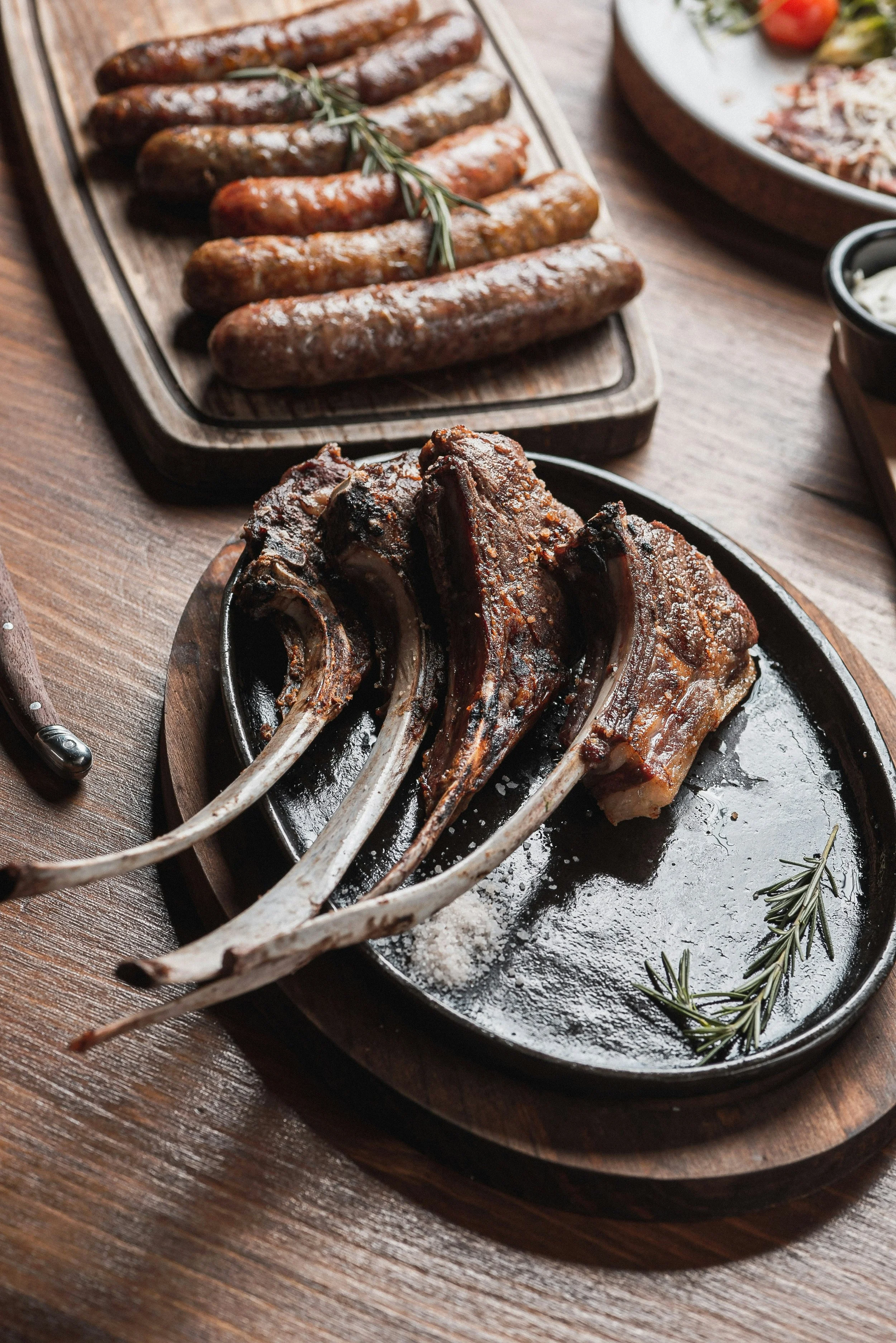 A black cast iron skillet with cooked lamb chops garnished with rosemary, placed on a round wooden serving board on a wooden table.