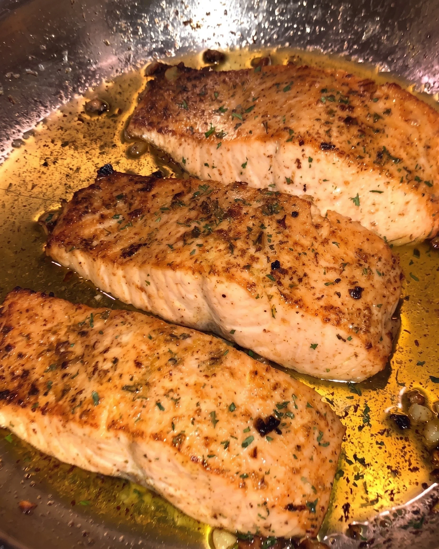 Tilapia fillets being cooked in a pan with seasonings and herbs, with some of the fish visible and a slightly browned exterior.