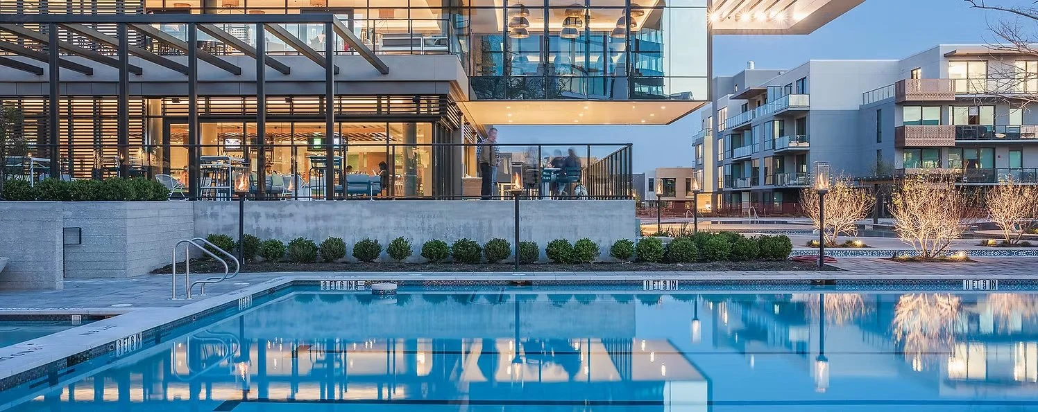 Modern apartment complex with outdoor pool, lit trees, and a restaurant or lounge area with people inside.