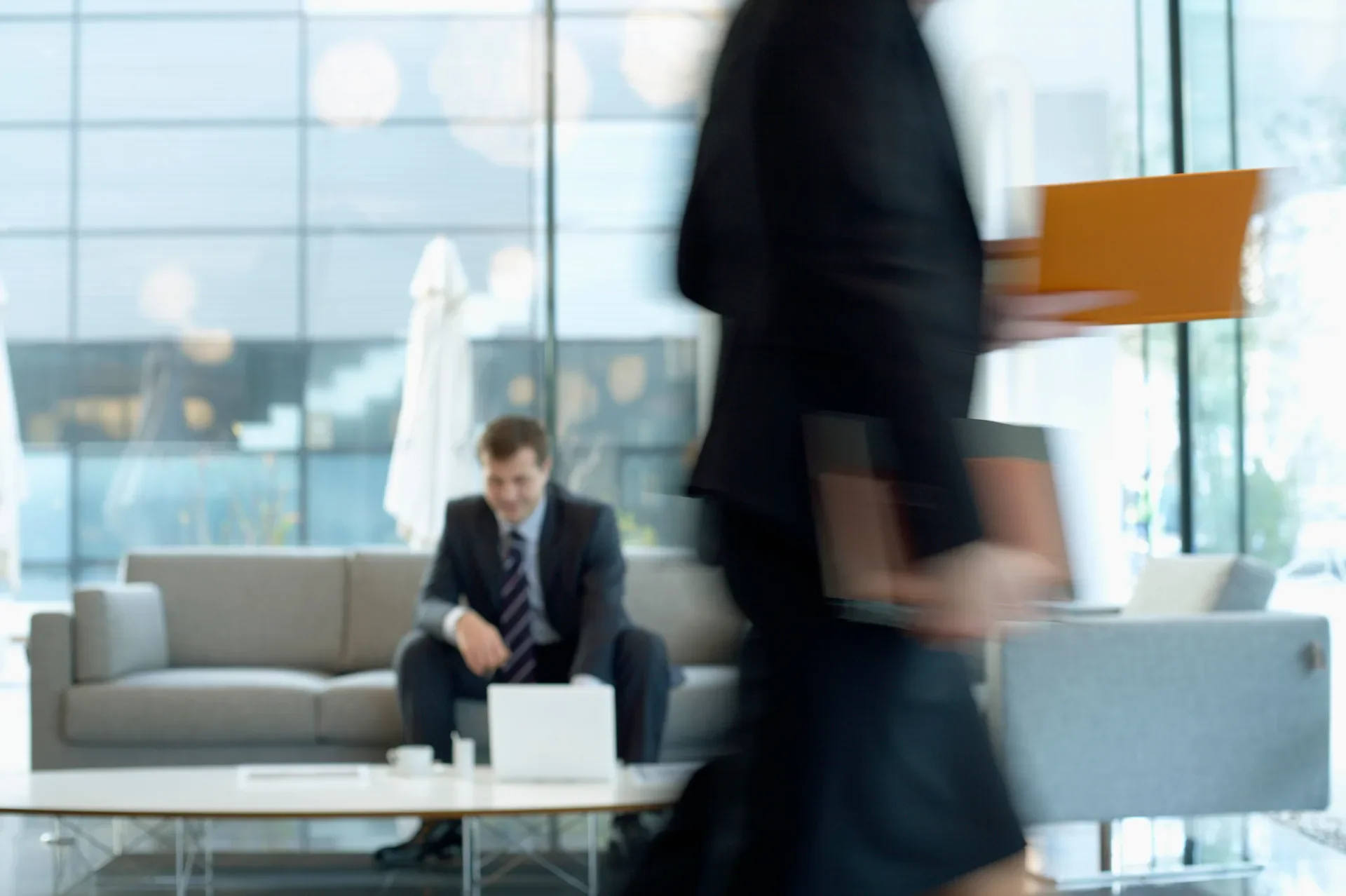 Businessman seated with a laptop in an office lobby as a colleague walks past in motion blur