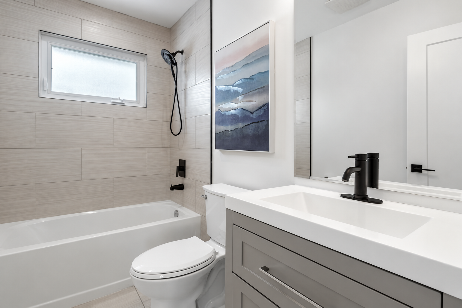 Modern bathroom with beige tiled walls, a bathtub, a white toilet, and a gray vanity with a white sink and black faucet. A large mirror, an abstract landscape painting, and a small frosted window are visible.