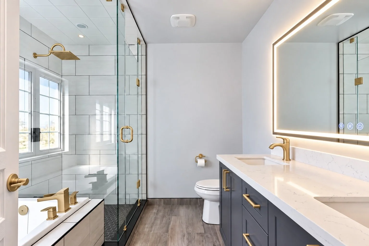 Modern bathroom with a glass shower enclosure, a bathtub with gold fixtures, a white toilet, a navy blue vanity with gold handles, a white marble countertop, and a large illuminated mirror.