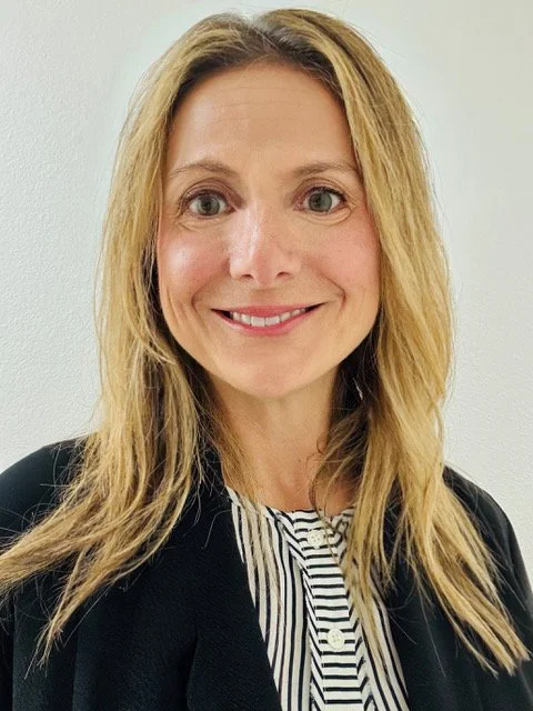 A woman with blonde hair smiling at the camera, wearing a black blazer over a black and white striped shirt, standing against a plain light-colored background.