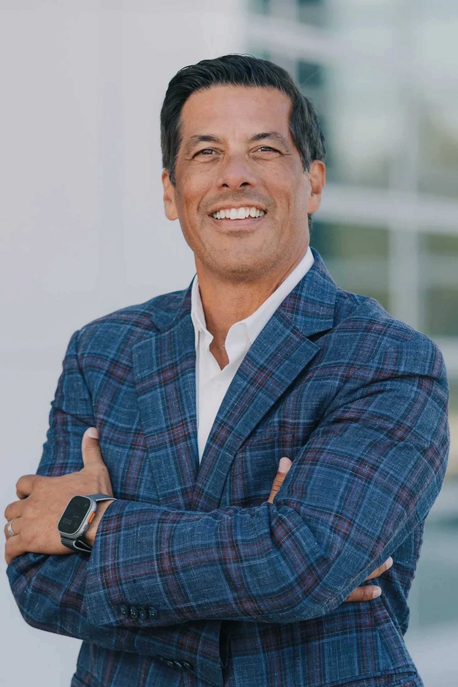 A middle-aged man with dark hair, wearing a checkered blue blazer, white shirt, and smartwatch, smiling confidently with arms crossed.