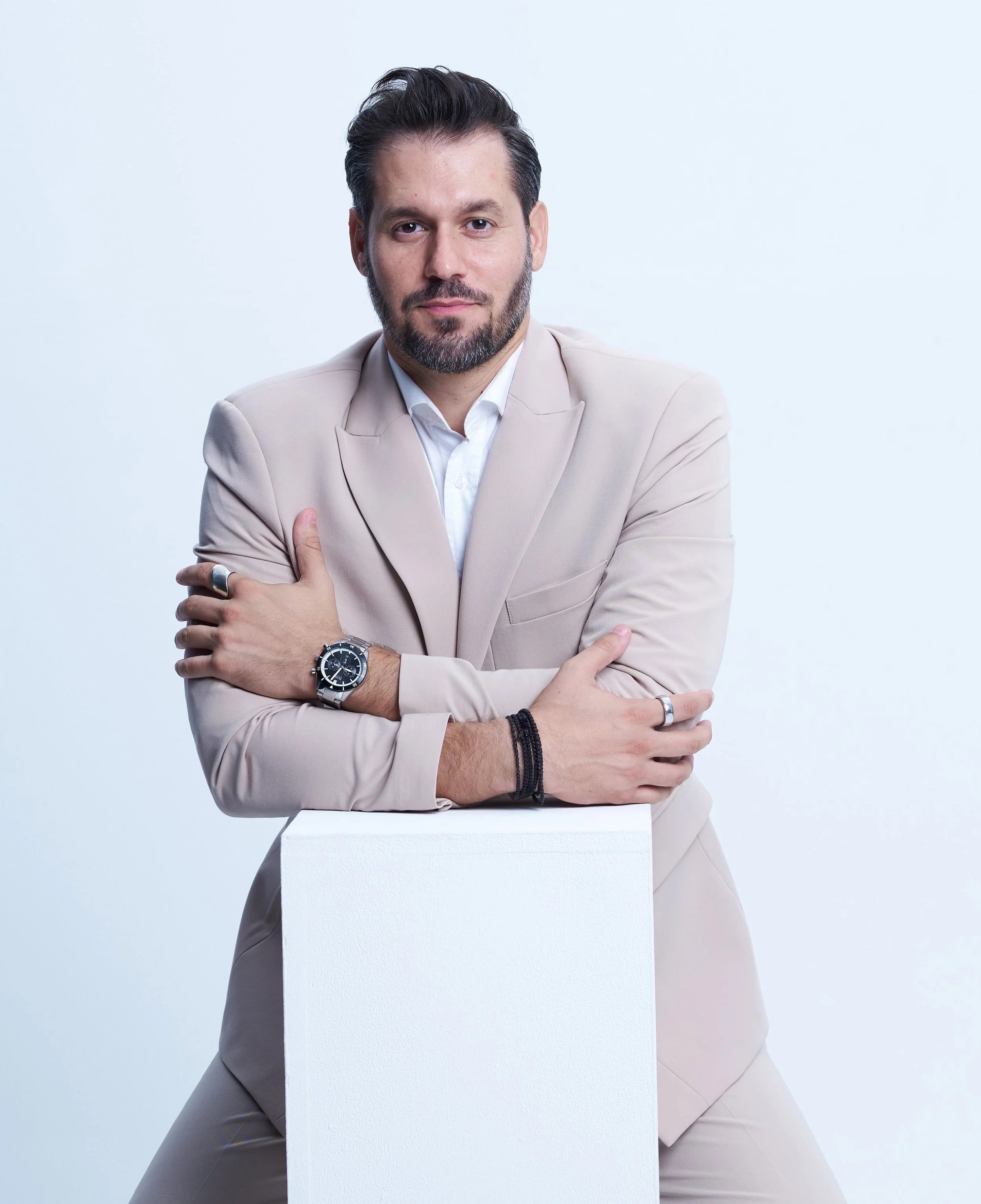 Man in a light beige suit with dark hair and beard, crossing arms, wearing watch and rings, standing behind a white pedestal on a light background.