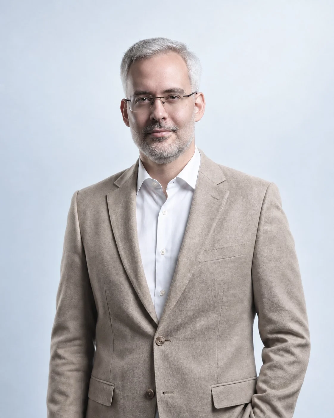 A middle-aged man with gray hair and glasses wearing a beige blazer and white shirt stands against a light gray background.