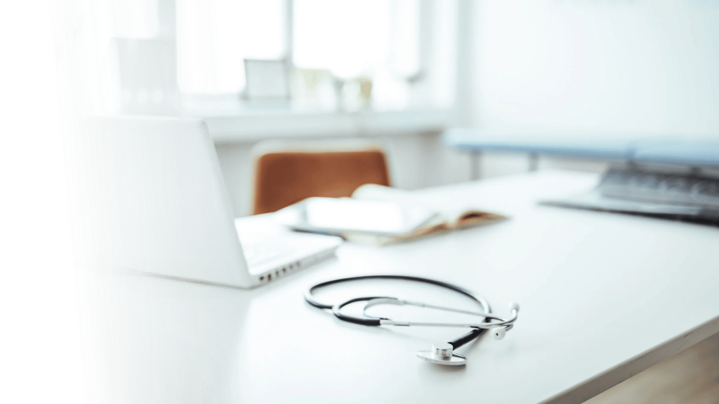 A stethoscope placed on a white desk in a well-lit medical or office setting, with a laptop and open book on the desk, background windows, and chairs.