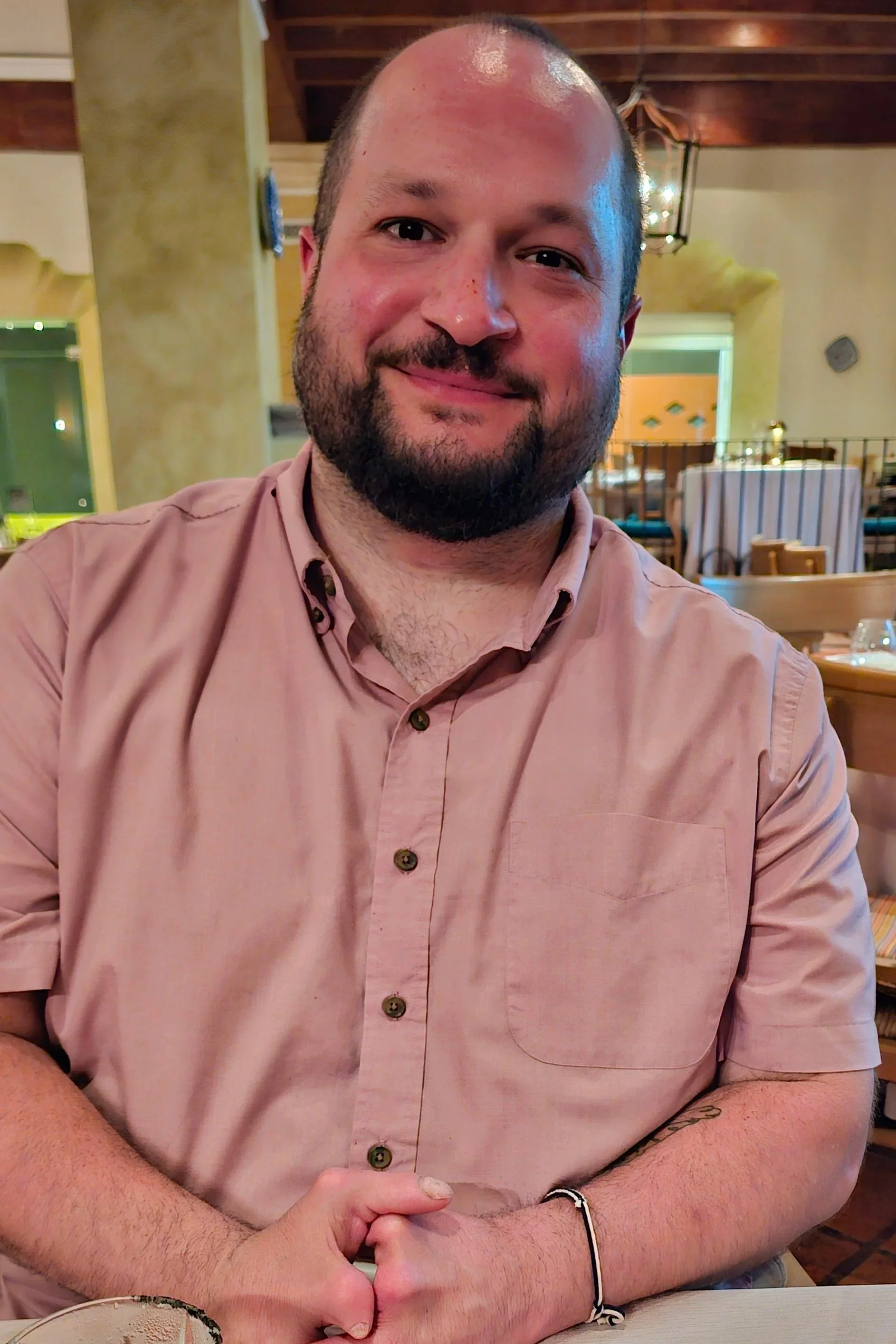 A man with a beard and short hair, wearing a pink shirt, smiling and sitting at a restaurant table with his hands clasped.