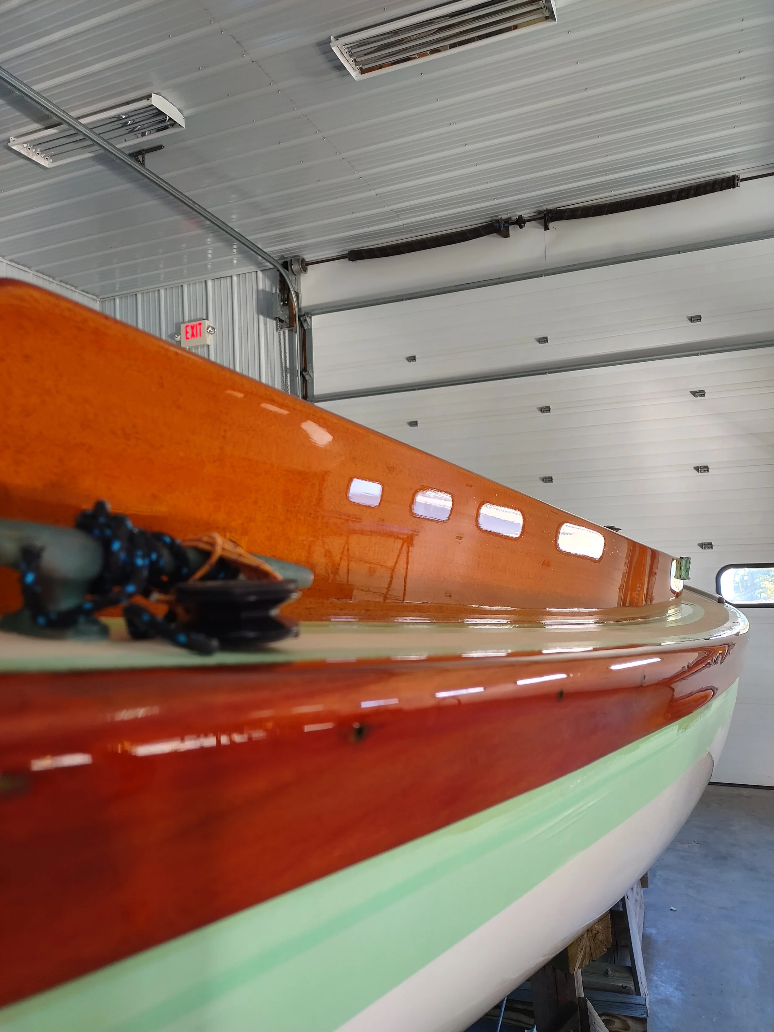 Close-up of the brightwork of a boat with a wooden rail, white bottom, and green stripe, inside a boat shop with a metallic ceiling.