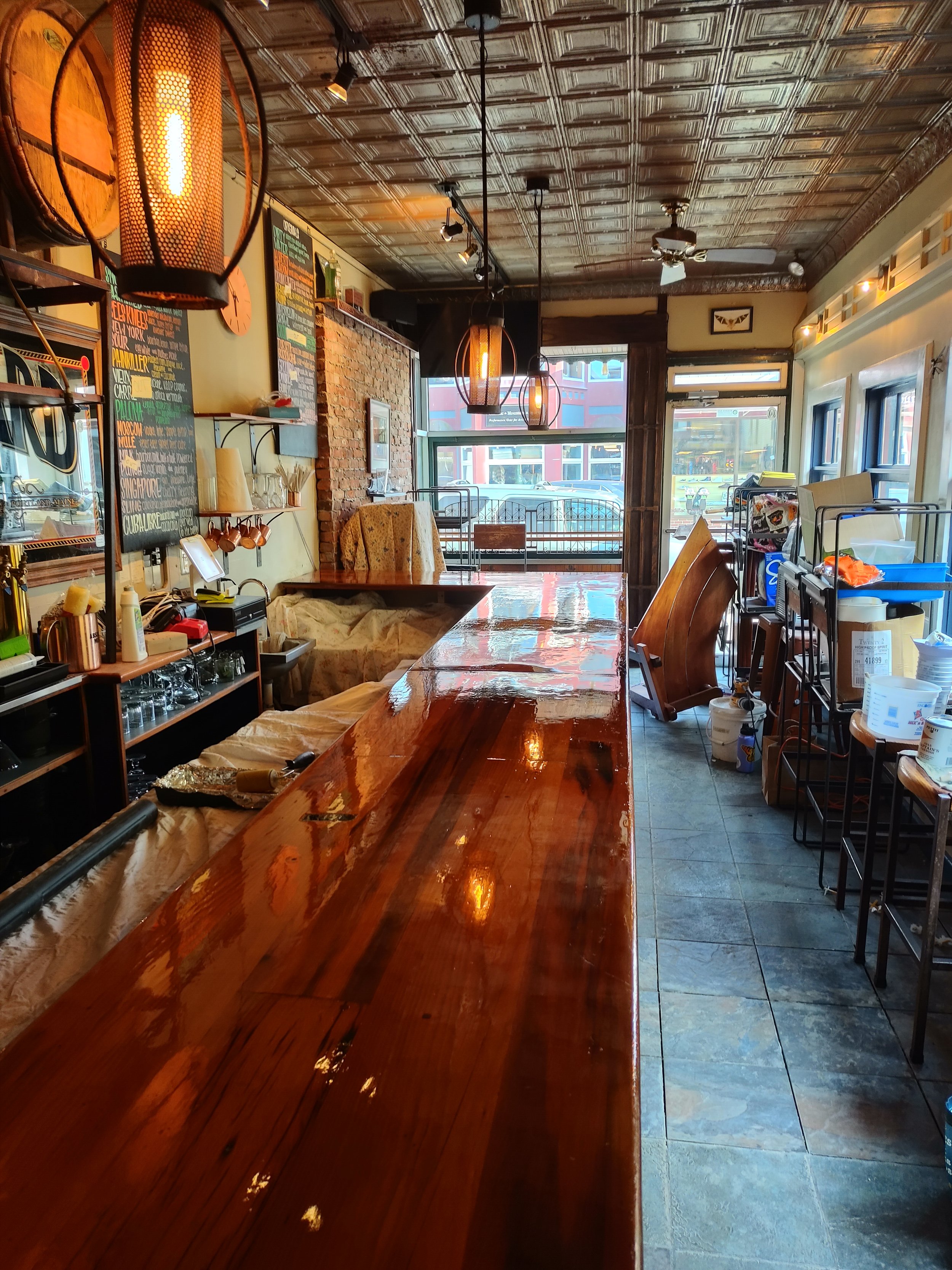 Empty restaurant interior with wooden bar counter, hanging light fixtures, brick and window walls, kitchen area with shelves and equipment, and a view outside through the windows.