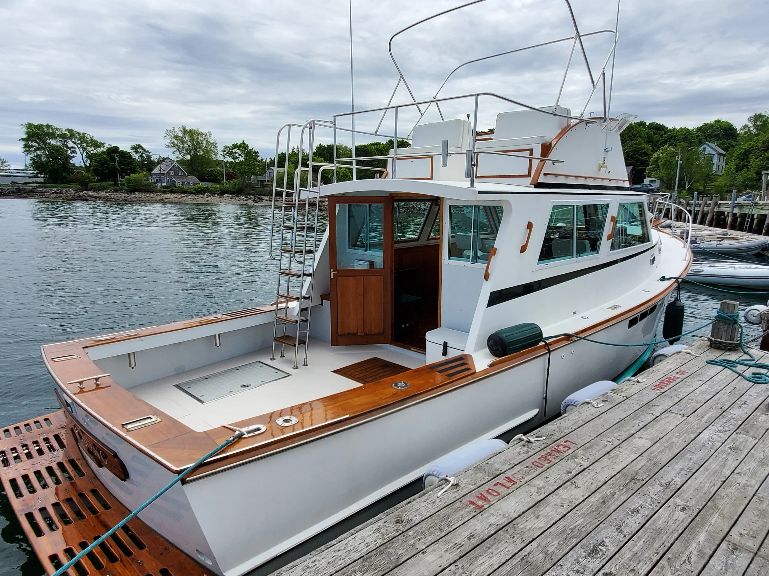 A white yacht docked at a wooden pier, with a cloudy sky and green trees in the background.
