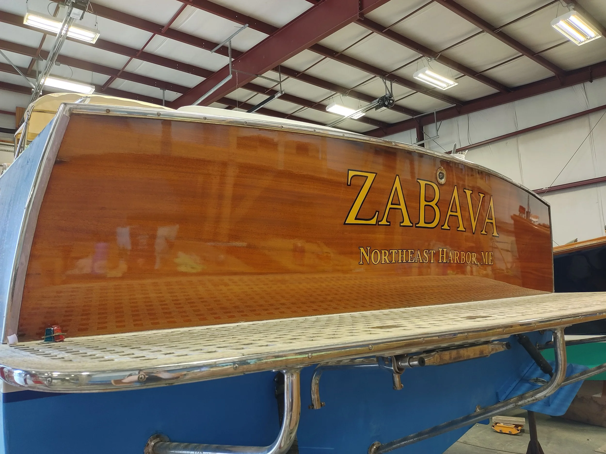 A wooden boat with the name 'ZABAVA' painted on the side, stored indoors with a metal scaffolding and a conveyor belt underneath.