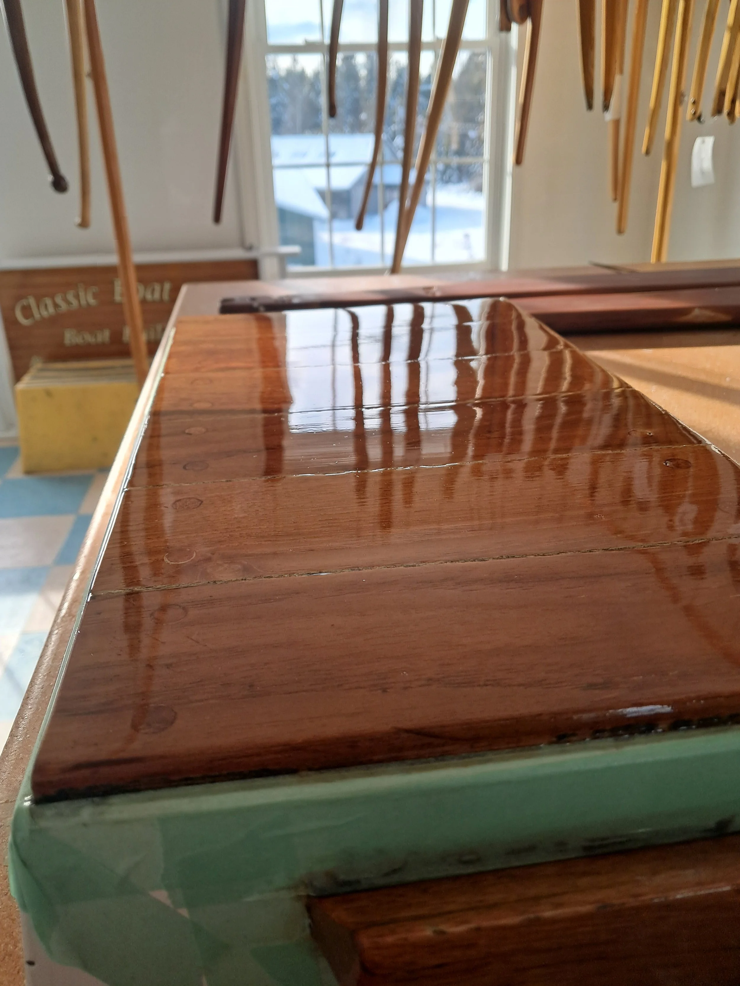 varnished boat parts with a painted green side and a shiny wooden top, inside a room with a window showing snowy trees outside.