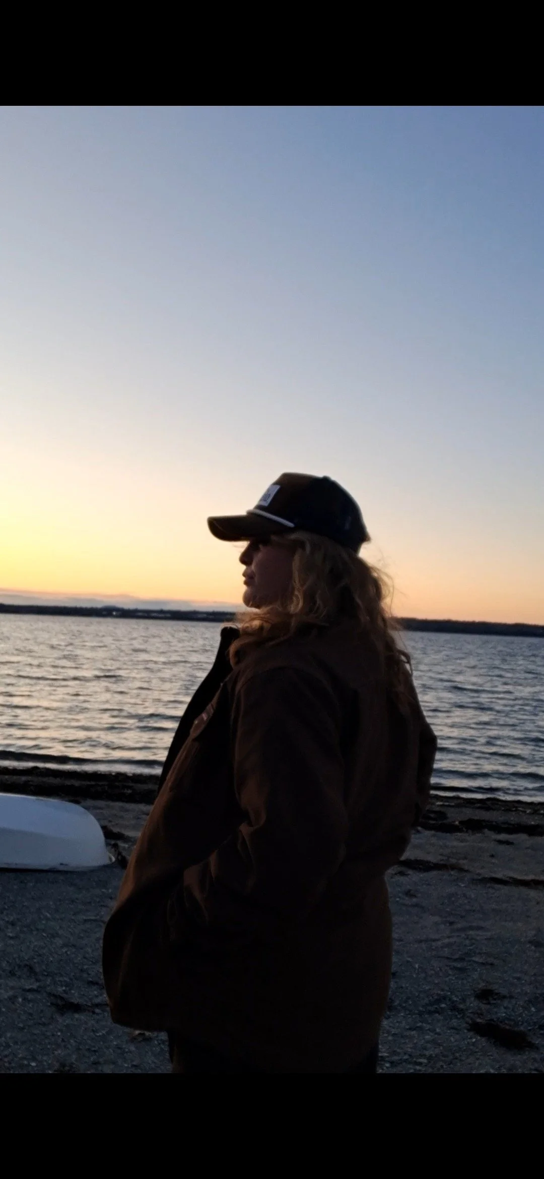 A woman in a brown jacket and a black baseball cap standing by a body of water during sunset.