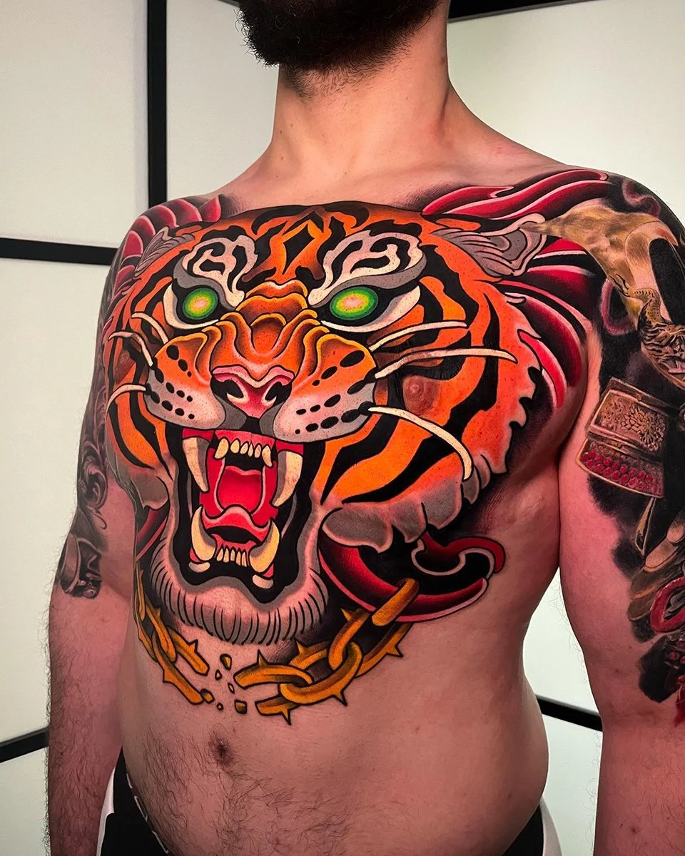 A person's chest and shoulder tattoo of a fierce, colorful tiger with green eyes, snarling with sharp teeth and a chain around its neck. By Johnny Domus, Neo Traditional and Neo Japanese Tattoo.