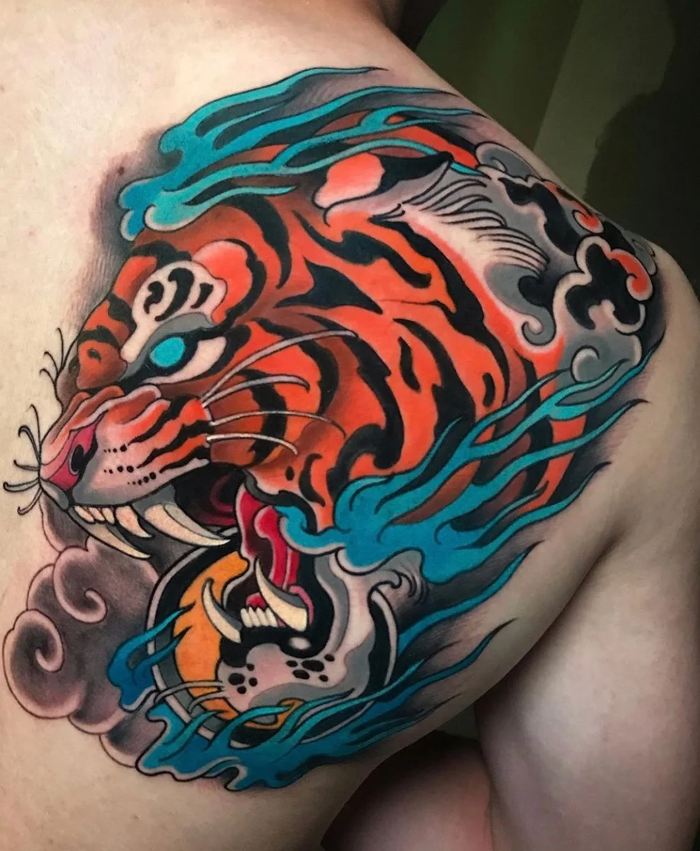Colorful tattoo of a tiger's face on a person's shoulder and chest with stylized waves in blue, black, and gray surrounding it. By Johnny Domus, Neo Traditional and Neo Japanese Tattoo.