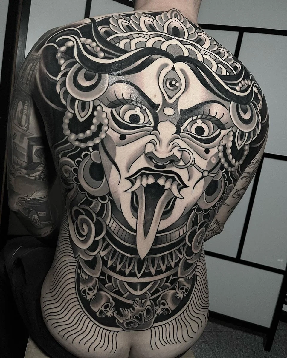 Black and gray tattoo of a fierce face with multiple eyes, fangs, and tongue on a person's back, featuring intricate patterns, skulls, and jewelry details. By Johnny Domus, Neo Traditional and Neo Japanese Tattoo.