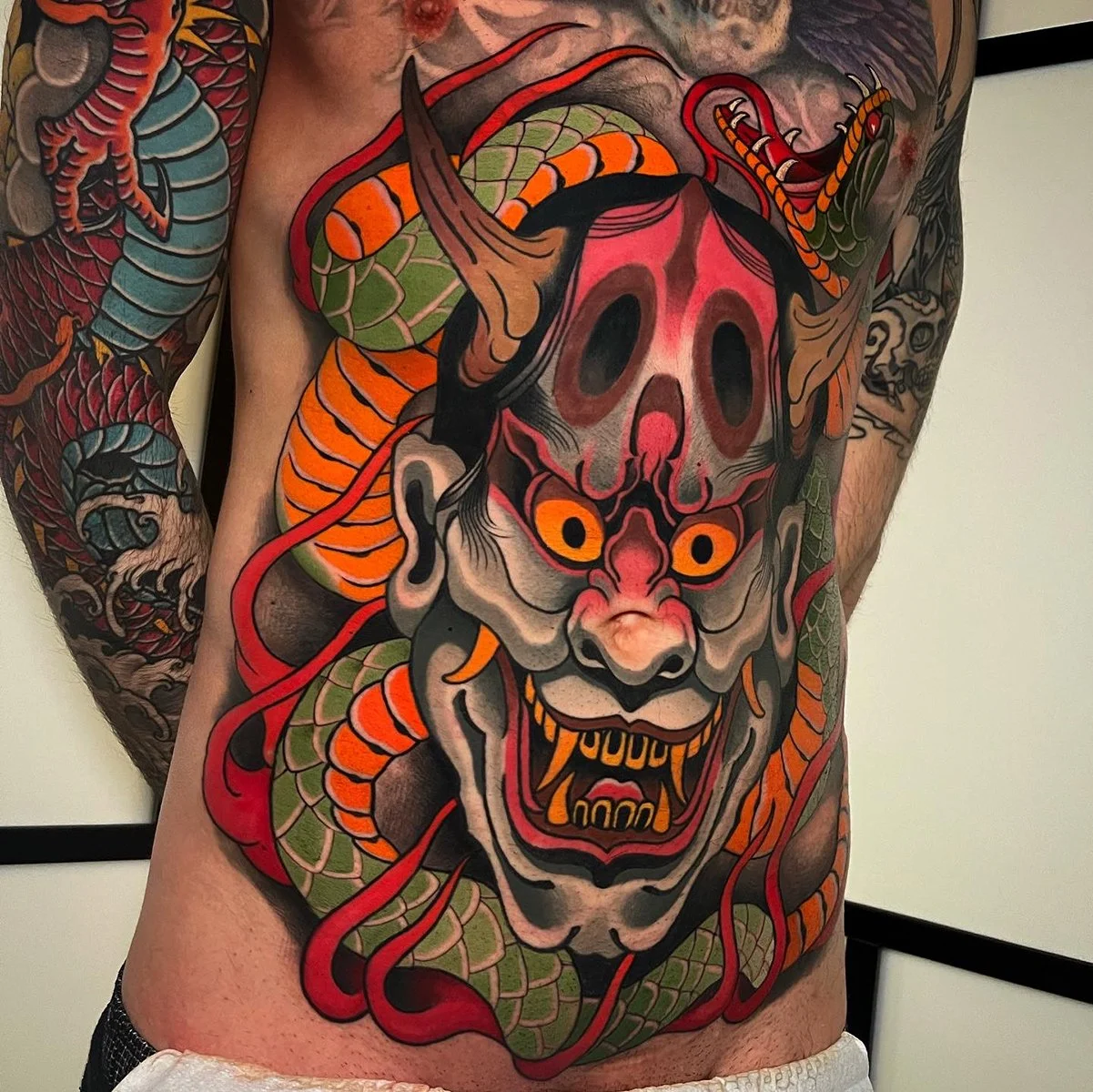 Tattoo of a fierce, colorful demon face with bright yellow eyes, sharp teeth, and a skull on top of its head, surrounded by red and green snakes on a person's chest. By Johnny Domus, Neo Traditional and Neo Japanese Tattoo.