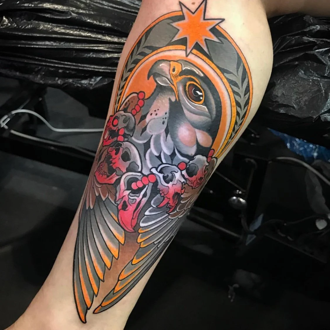 Tattoo of a falcon with a radiant star, surrounded by decorative elements in orange, red, black, and gray ink on a person's forearm. By Johnny Domus, Neo Traditional and Neo Japanese Tattoo.