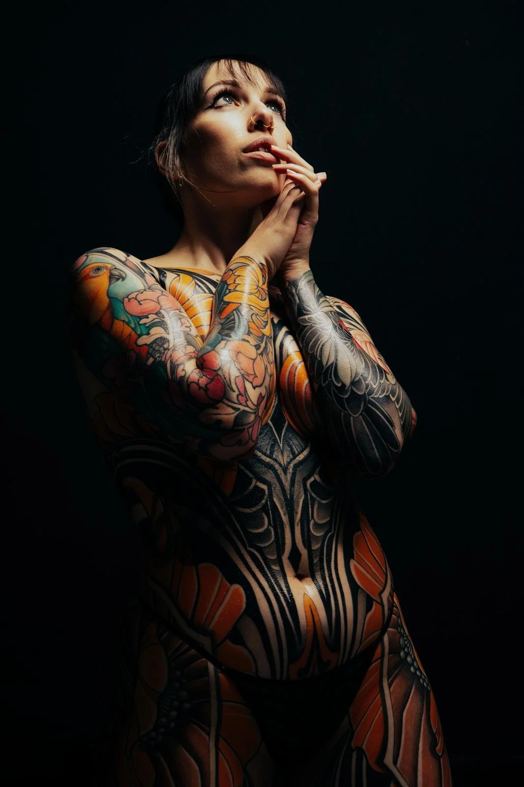 Tattooed woman with dark hair and bangs, looking upward with hands touching her face, against black background.