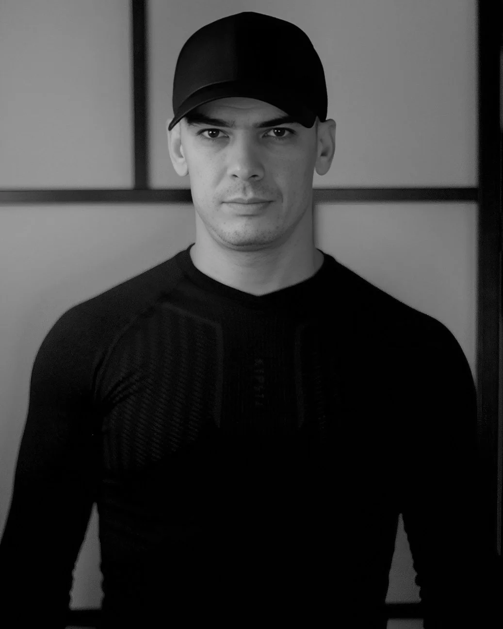 Black and white portrait of a young man wearing a cap and athletic shirt, standing indoors against a background with rectangular panels. By Johnny Domus, Neo Traditional and Neo Japanese Tattoo.
