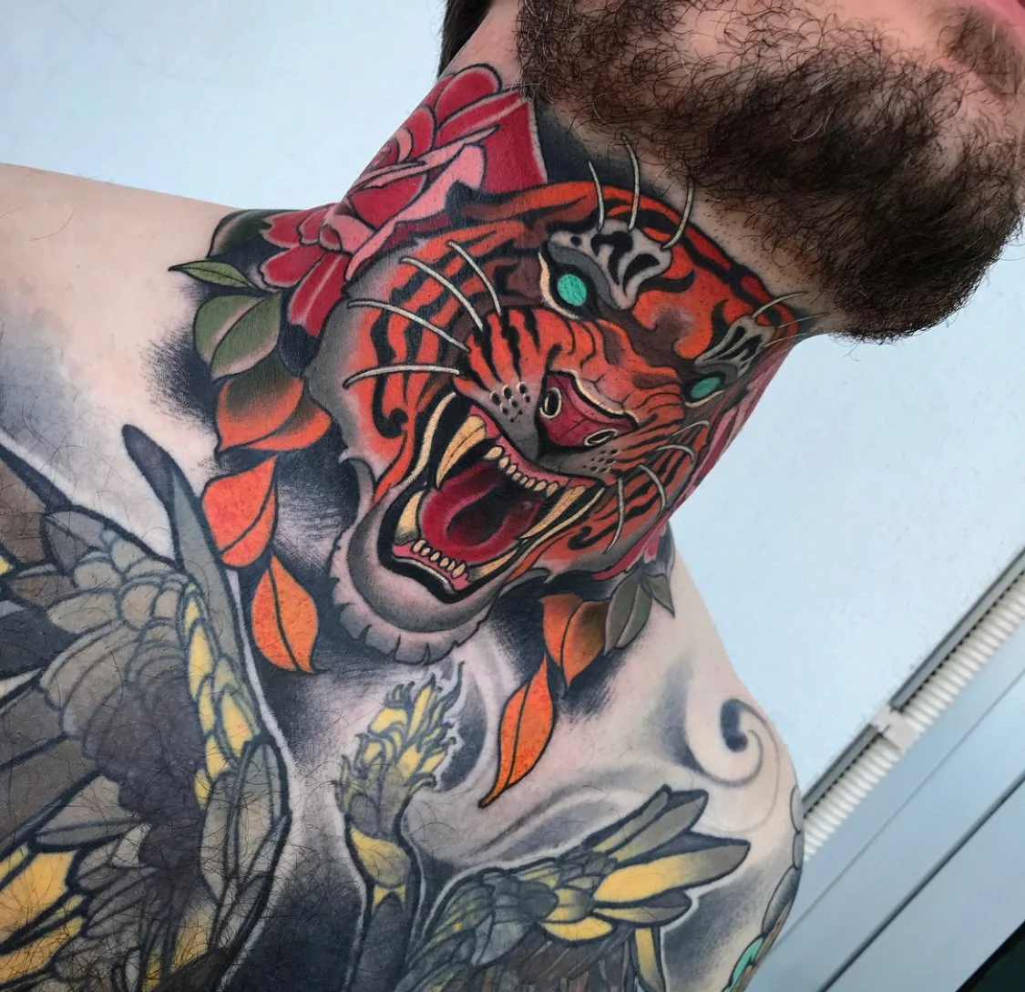 A close-up of a tattoo on a man's chest and neck, featuring a fierce, colorful tiger's head with its mouth open, surrounded by floral and leaf designs. By Johnny Domus, Neo Traditional and Neo Japanese Tattoo.