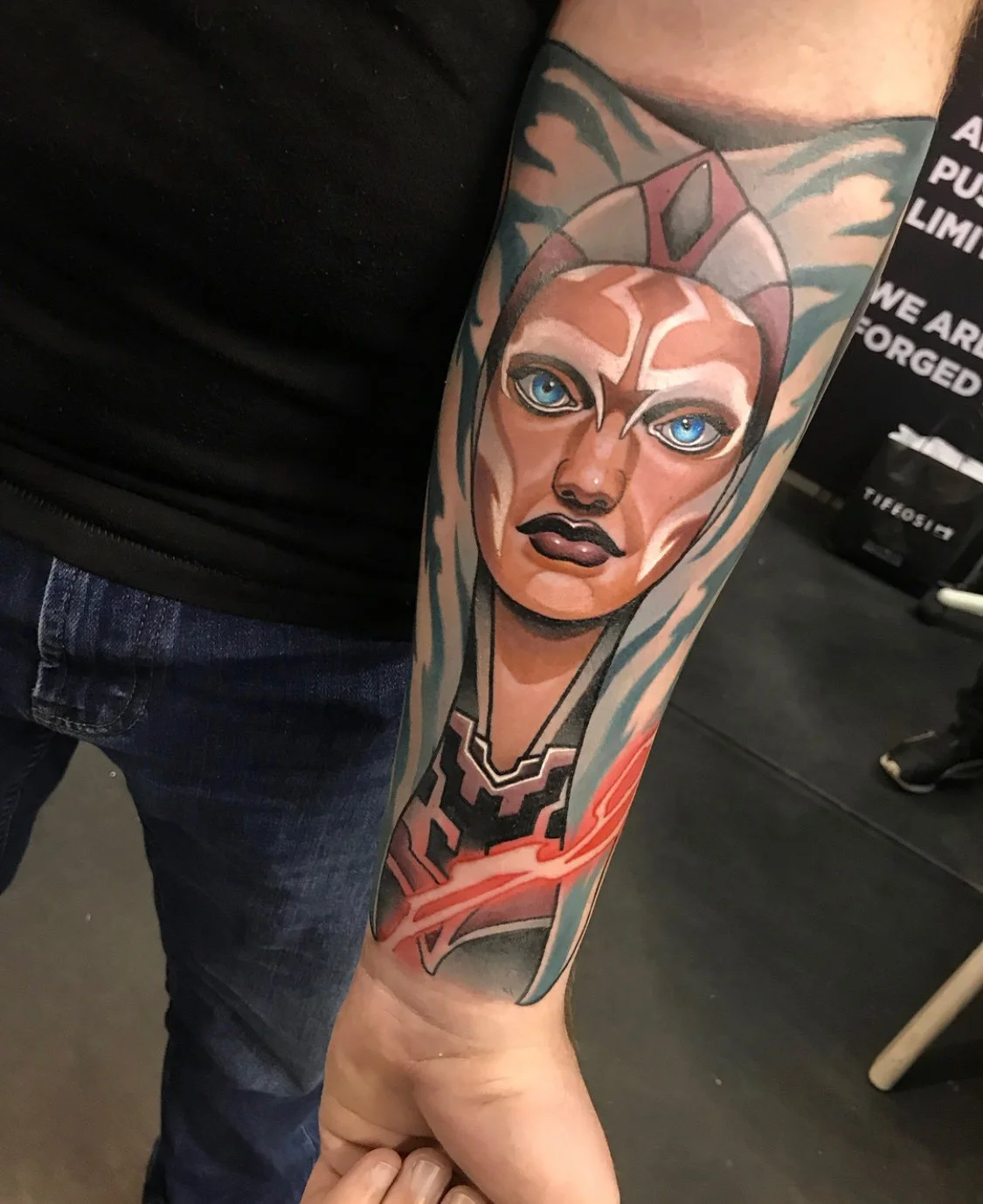 Colorful tattoo on a person's arm depicting a female face with blue eyes, dark lipstick, and a headdress, with a stylized design. By Johnny Domus, Neo Traditional and Neo Japanese Tattoo.