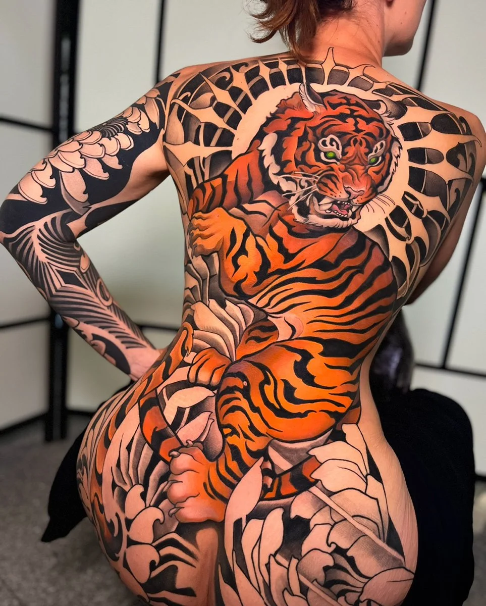 A person with extensive tiger-themed tattoos covering their chest, back, and arms, featuring a fierce orange and black tiger with green eyes. By Johnny Domus, Neo Traditional and Neo Japanese Tattoo.