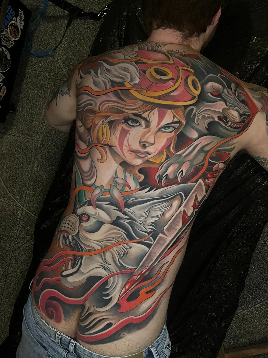 A man lying face down with a large, colorful tattoo covering his back. The tattoo features a woman's face with flowing hair, surrounded by mythical and fantasy elements such as a creature resembling a wolf or lion, flames, and abstract designs. By Jo