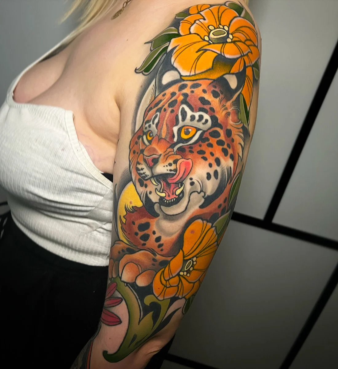Tattoo of a fierce tiger with orange fur, black spots, and yellow eyes on a person's upper arm, surrounded by orange and yellow flowers and green leaves. By Johnny Domus, Neo Traditional and Neo Japanese Tattoo.