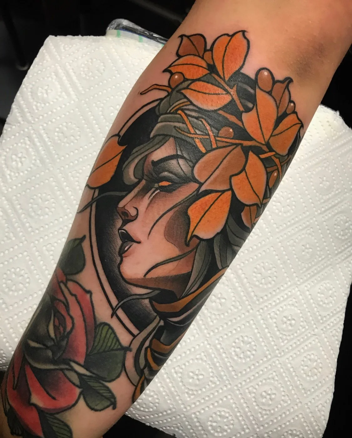 Tattoo of a woman's face with closed eyes, surrounded by orange leaves and a red rose on a person's forearm. By Johnny Domus, Neo Traditional and Neo Japanese Tattoo.