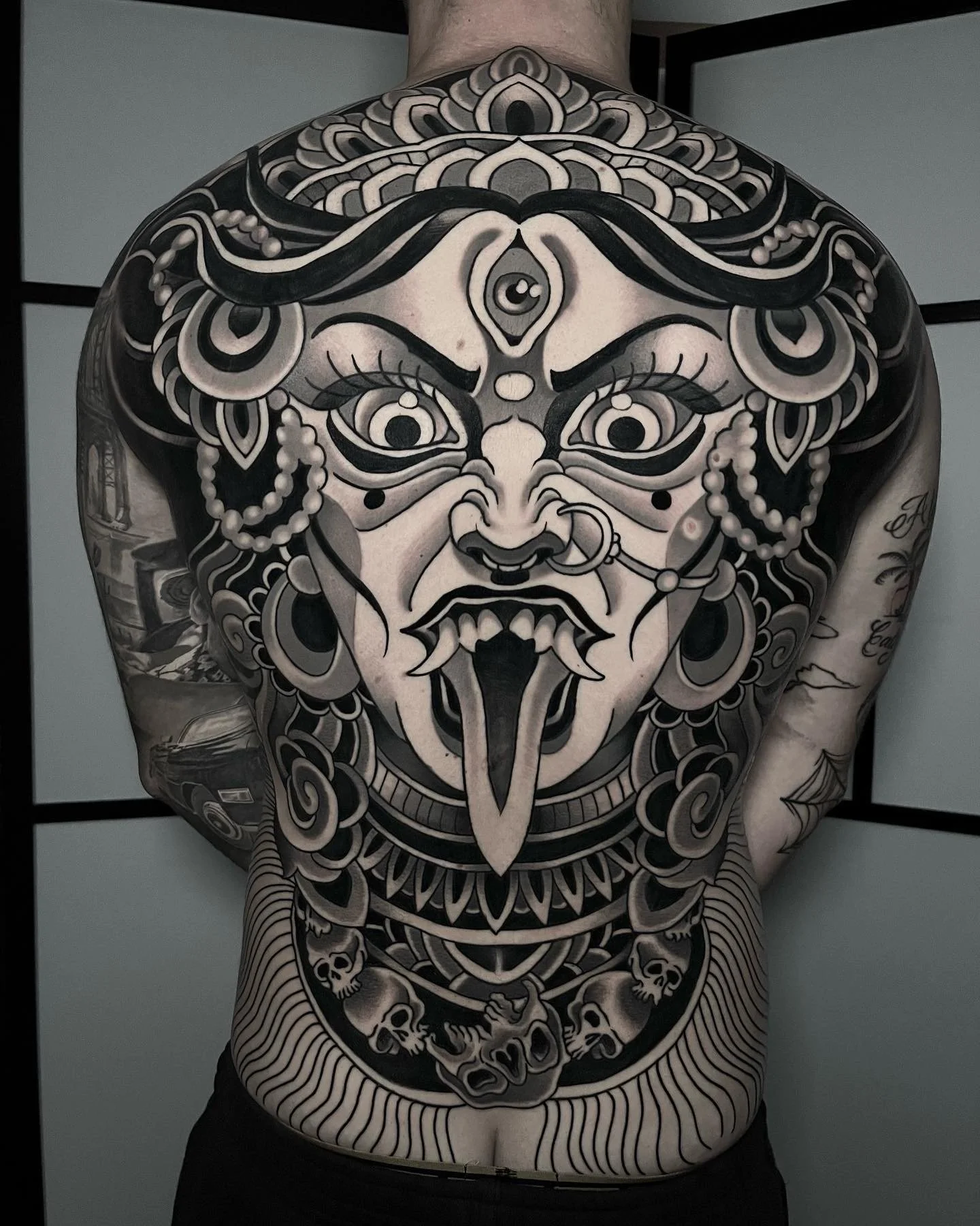 A black and white tattoo of a fierce deity face with multiple eyes, prominent eyebrows, and elaborate headpieces covering the upper back and shoulders, with additional skull and skull motif tattoos on the lower back and arms.