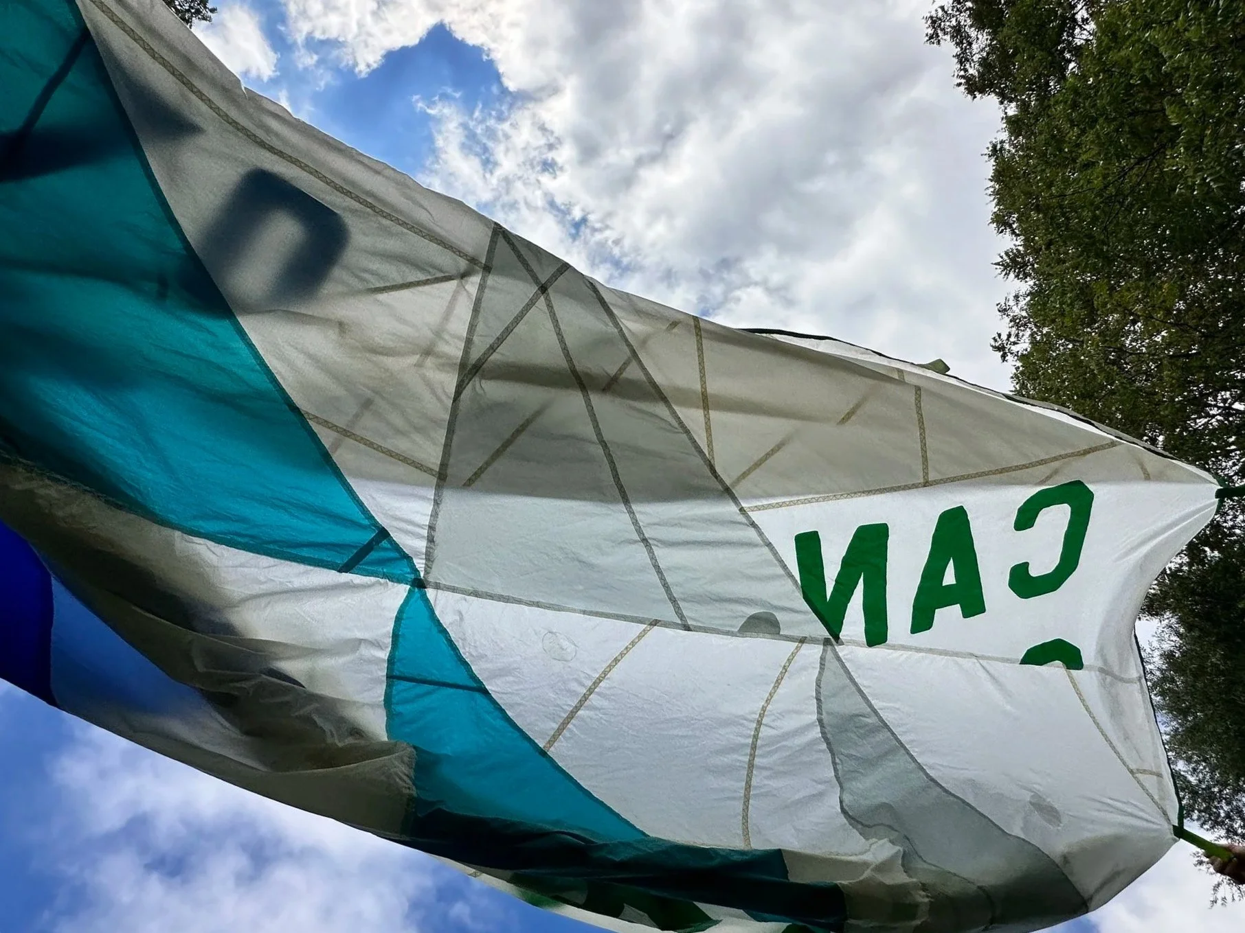 Across a cloudy sky is a piece of quilted fabric blowing across the image.  The fabric is quilted in large white, turquoise and grey triangles with some green lettering on the fabric.