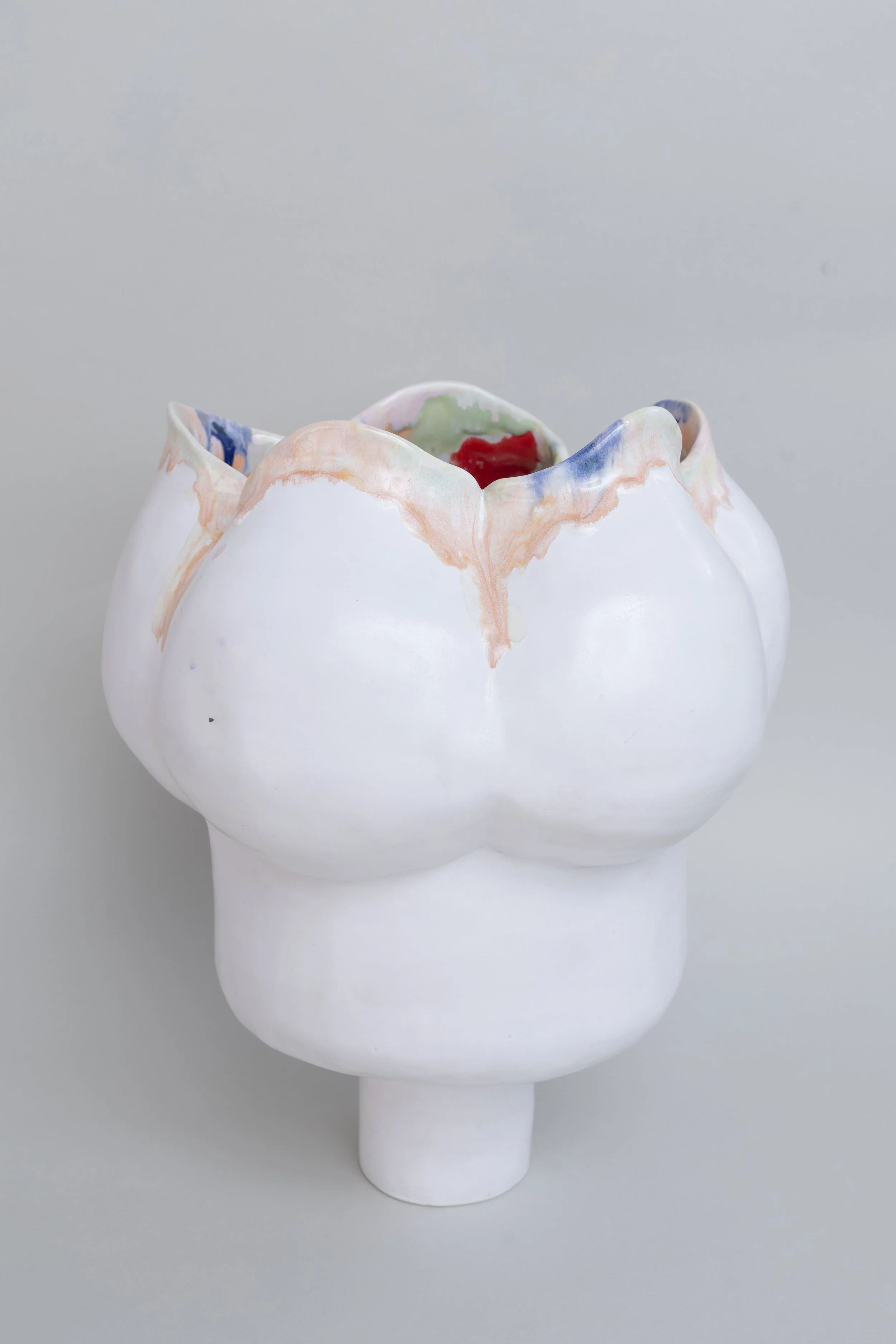 Ceramic sculpture with a circular foot and base billowing out into petals similar to a flower.  The outside is a matte white with drips of coloured glaze along the edge of the petals.  The inside of the vessel has various coloured glazes melting.