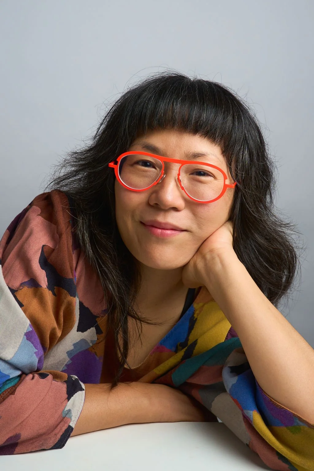 A woman with shoulder-length black hair and glasses with red frames, resting her chin on her hand and smiling, wearing a colorful patterned top, against a plain gray background.