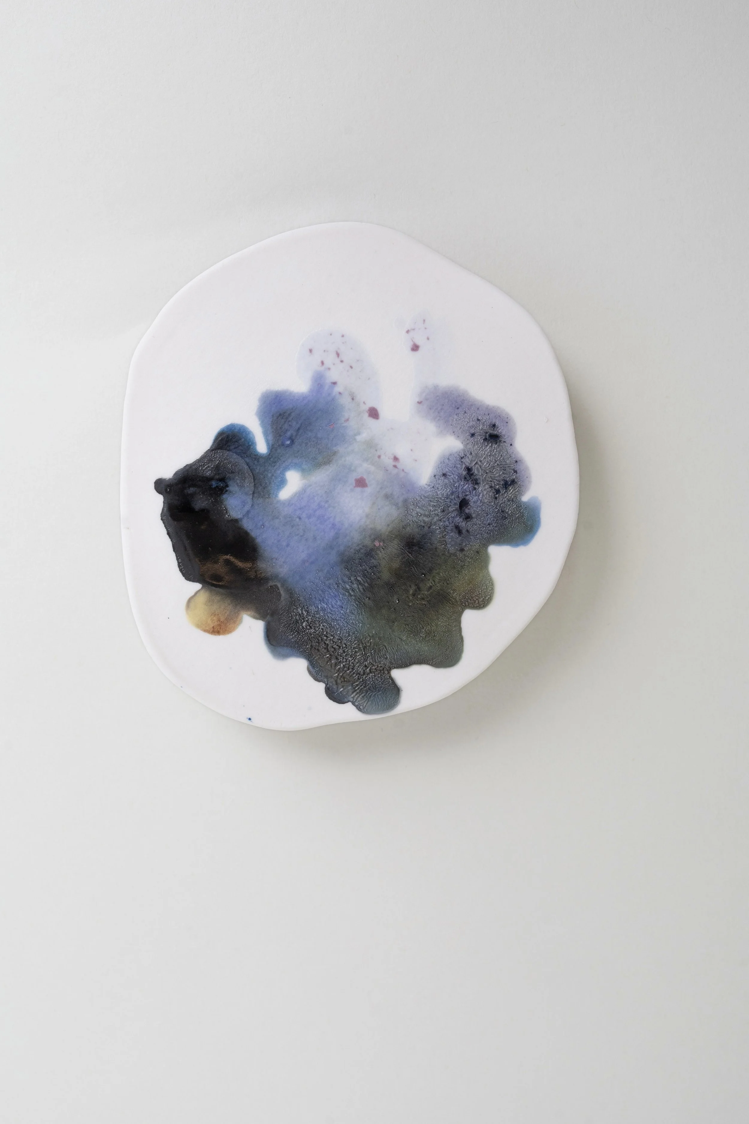 A hand rolled porcelain tile with black, dark blue and hints of green glaze smoothly blending into each other along one side of the glaze patch.  The other side of the glaze gently lightens to a lighter blue and then into a transparent purple.